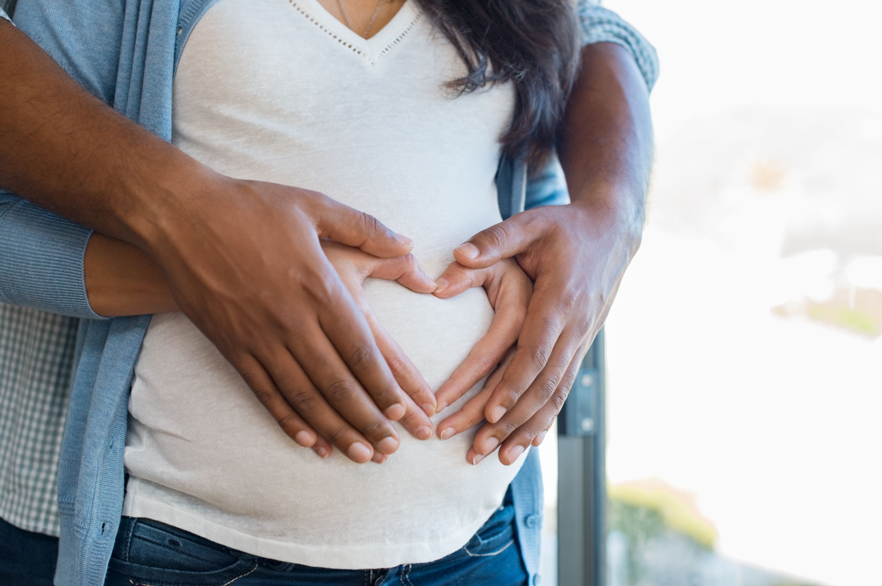 Husband and wife hold hands in a shape of a heart over pregnant wifes stomach